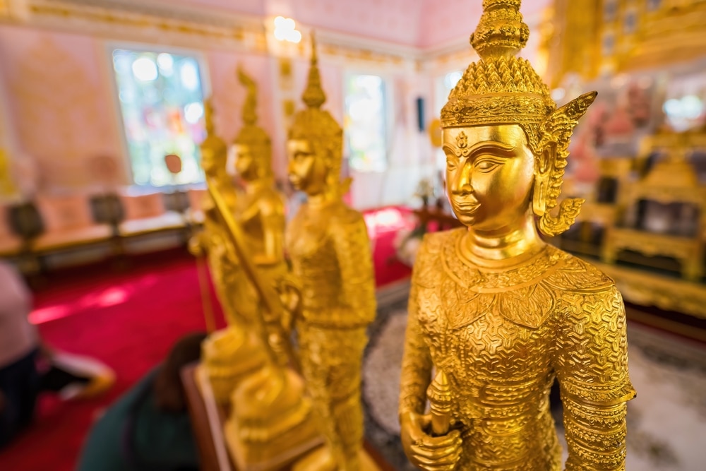 แถวของรูปปั้นสีทองสไตล์ไทยภายในอาคารที่ตกแต่งอย่างวิจิตรภายในสถานที่ท่องเที่ยวยอดนิยมในนครนายก.