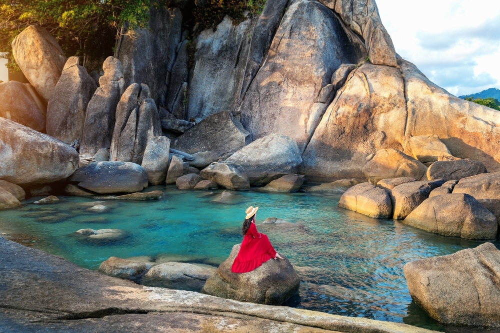 มีผู้หญิงคนหนึ่งสวมชุดสีแดงและหมวกฟาง นั่งอยู่บนก้อนหินในสระน้ำทะเลสีเทอร์ควอยซ์ ที่ล้อมรอบด้วยก้อนหินแกรนิต เป็นสถานที่ที่สวยงามสำหรับผู้ที่เดินทาง ที่เที่ยวเกาะสมุย 2026 