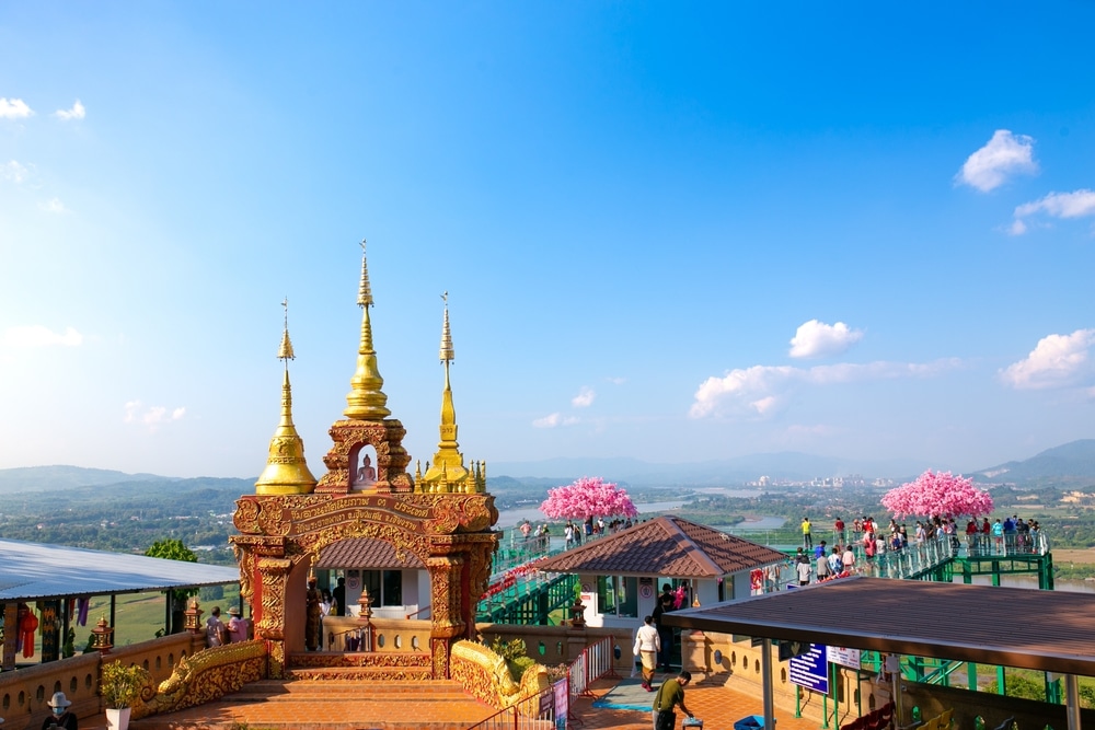 ประตูวัดที่ตกแต่งอย่างวิจิตรงดงามสีทอง ตั้งอยู่บนดาดฟ้าชมวิวที่มีพื้นกระจก พร้อมด้วยต้นซากุระสีชมพูเทียม ซึ่งมองเห็นหุบเขาแม่น้ำได้อย่างชัดเจน เป็นสถานที่ท่องเที่ยวยอดนิยมในเชียงราย.