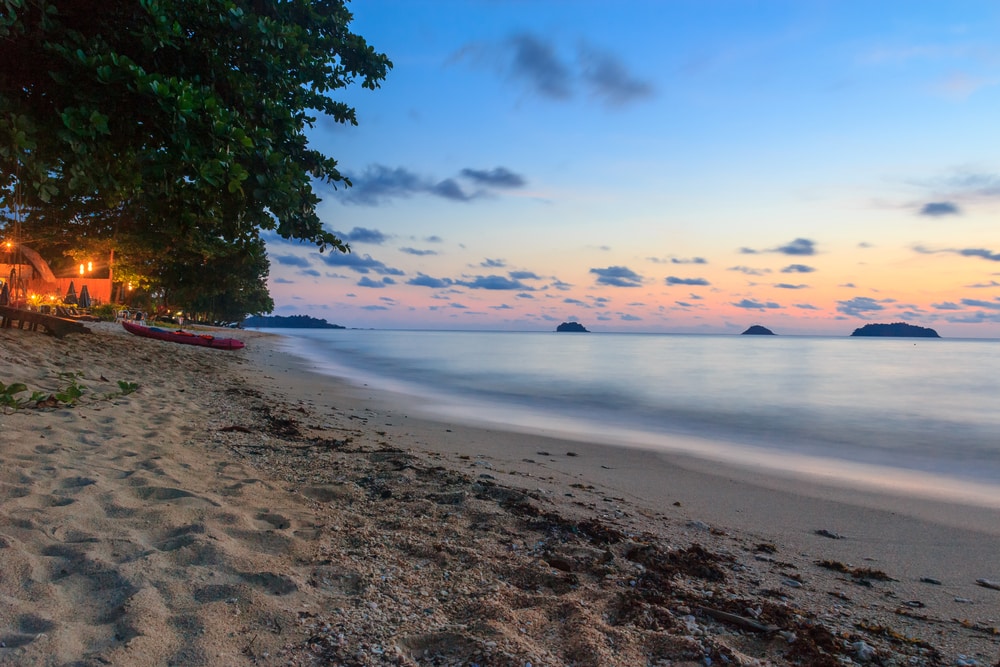 ชายหาดทรายในยามพลบค่ำ น้ำทะเลสงบ มีเกาะเล็ก ๆ อยู่ห่างจากฝั่ง และต้นไม้เรียงรายตามแนวชายฝั่ง ใกล้ที่พักราคาไม่แพงบนเกาะลัน.