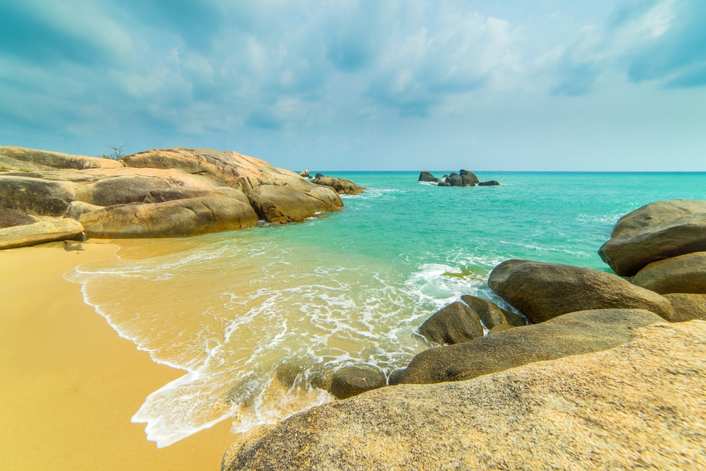 น้ำสีเทอร์ควอยซ์ซัดเข้าหาชายหาดทรายที่ล้อมรอบด้วยหินก้อนใหญ่เรียบเนียน ใต้ท้องฟ้าสีฟ้าที่มีเมฆครึ้ม เป็นจุดชมวิวที่สวยงามสำหรับผู้ที่เดินทาง ที่เที่ยวเกาะสมุย 2026 