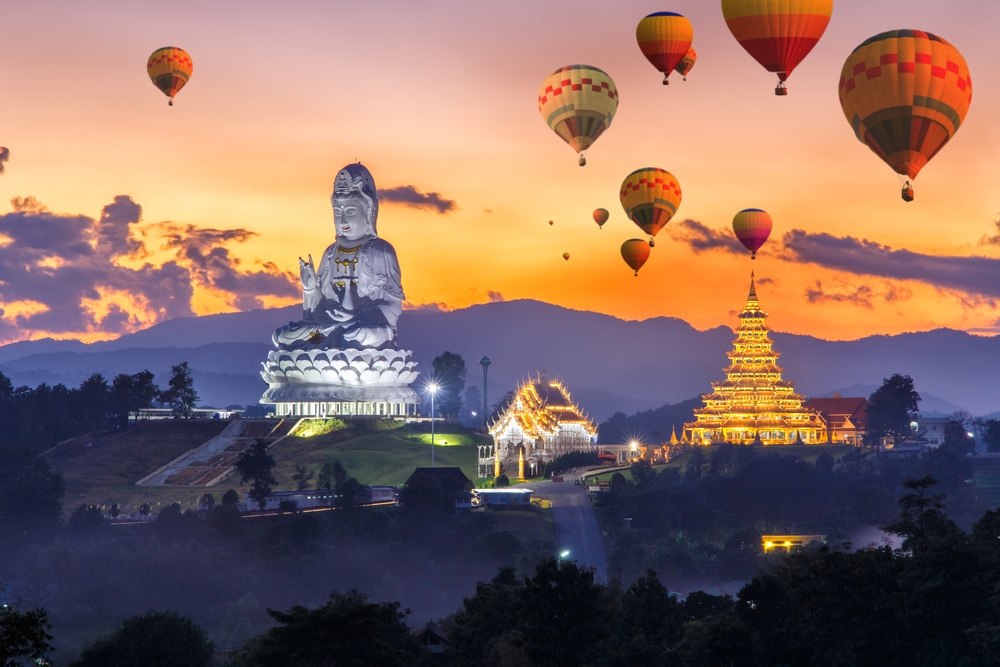 บอลลูนอากาศร้อนลอยอยู่เหนือพระพุทธรูปสีขาวขนาดใหญ่และกลุ่มวัดสีทองในช่วงพระอาทิตย์ตกดิน ซึ่งเป็นจุดเด่นสำหรับผู้ที่มาเที่ยวเชียงราย.
