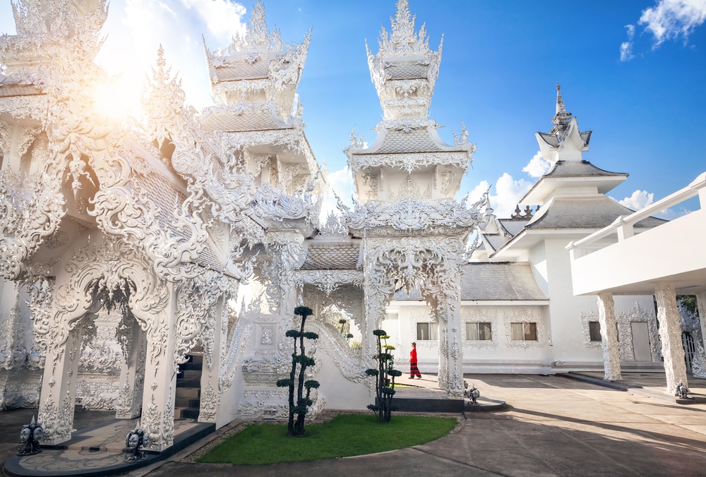 อาคารวัดสีขาวที่สลักลวดลายอย่างประณีต พร้อมด้วยหลังคาหลายชั้นที่วัดร่องขุน ซึ่งเป็นสถานที่ท่องเที่ยวยอดนิยมในเชียงราย ขณะที่ชายคนหนึ่งสวมเสื้อสีแดงกำลังเดินผ่านระหว่างอาคารเหล่านั้น.