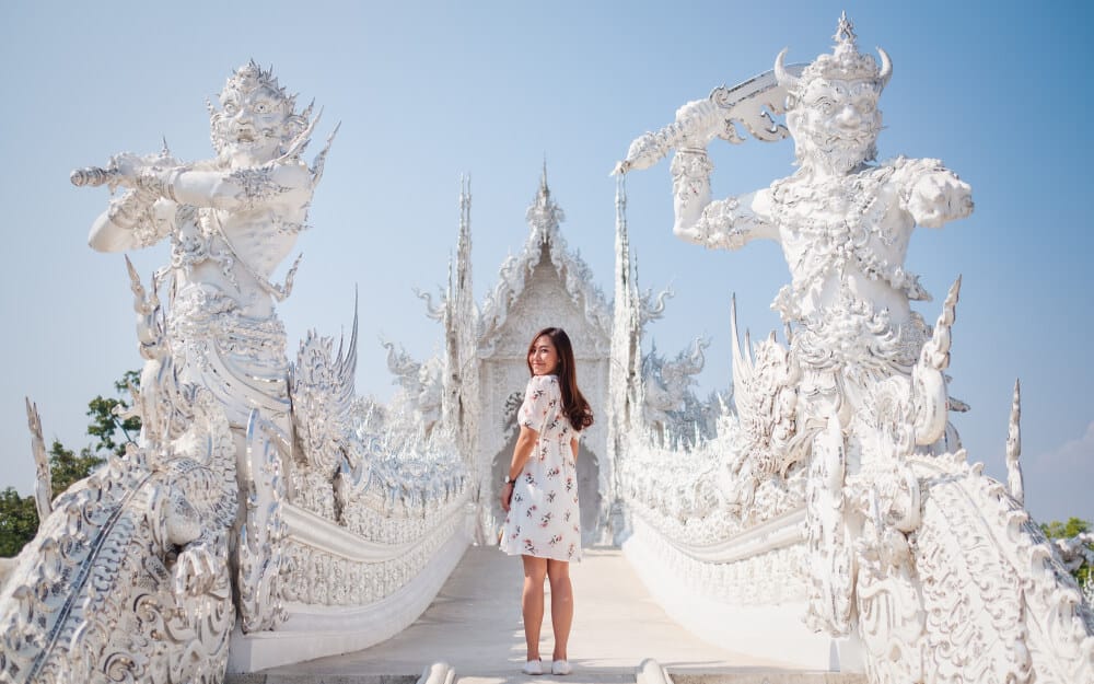 ผู้หญิงคนหนึ่งสวมชุดลายดอกยืนอยู่บนสะพานระหว่างรูปปั้นสีขาวที่ตกแต่งอย่างวิจิตรสององค์ ณ วัดแห่งหนึ่ง ขณะกำลังเยี่ยมชมสถานที่ท่องเที่ยวชั้นนำในเชียงราย.