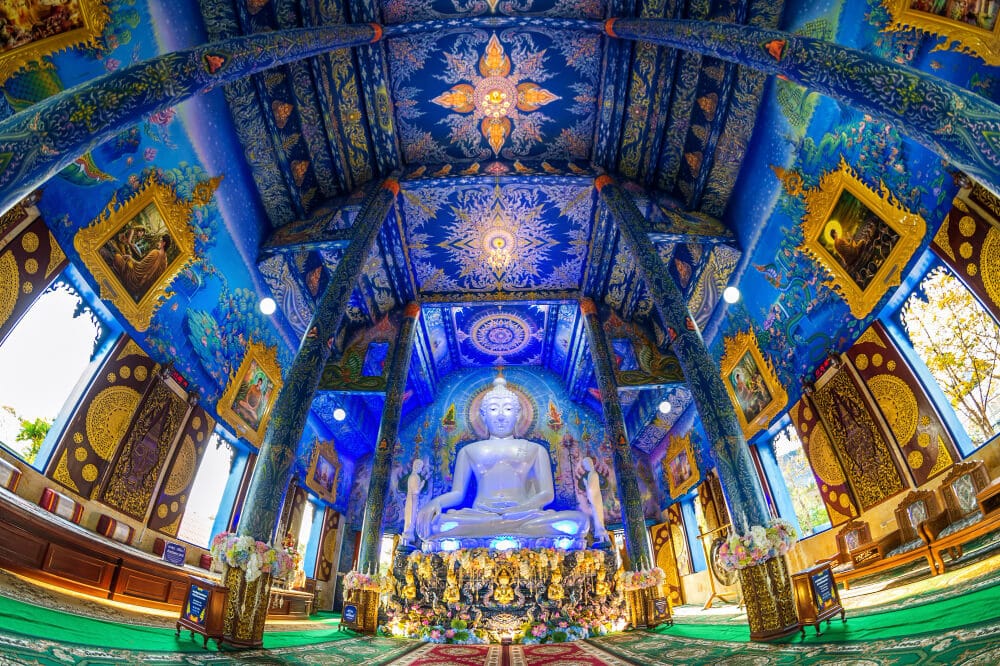 ภายในวัดในจังหวัดเชียงราย มีผนังและเพดานสีน้ำเงินเข้ม พร้อมด้วยลวดลายที่ซับซ้อนล้อมรอบพระพุทธรูปสีขาวขนาดใหญ่ที่ตั้งอยู่บนฐานทอง.