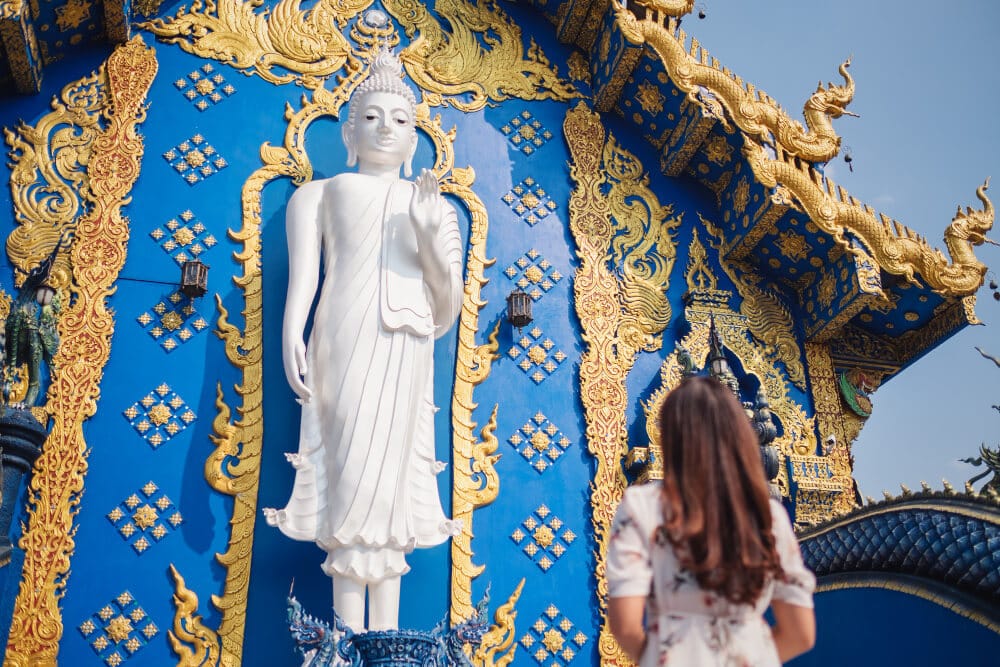 ผู้หญิงคนหนึ่งยืนอยู่หน้ารูปปั้นพระพุทธรูปสีขาวขนาดใหญ่ บนพื้นหลังของวัดร่องเสี้ยวเท็นที่มีผนังด้านนอกสีฟ้าและสีทองประดับตกแต่งอย่างวิจิตร ซึ่งเป็นจุดหมายปลายทางที่ไม่ควรพลาดสำหรับผู้ที่ชื่นชอบการเที่ยวเชียงราย.